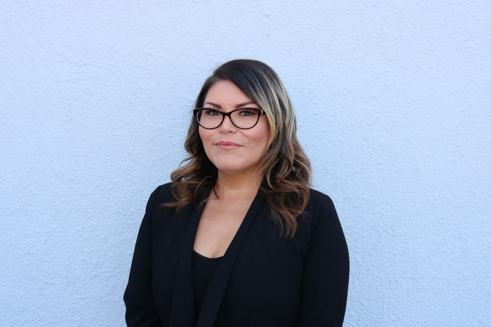 A woman in glasses and black jacket standing next to blue wall.